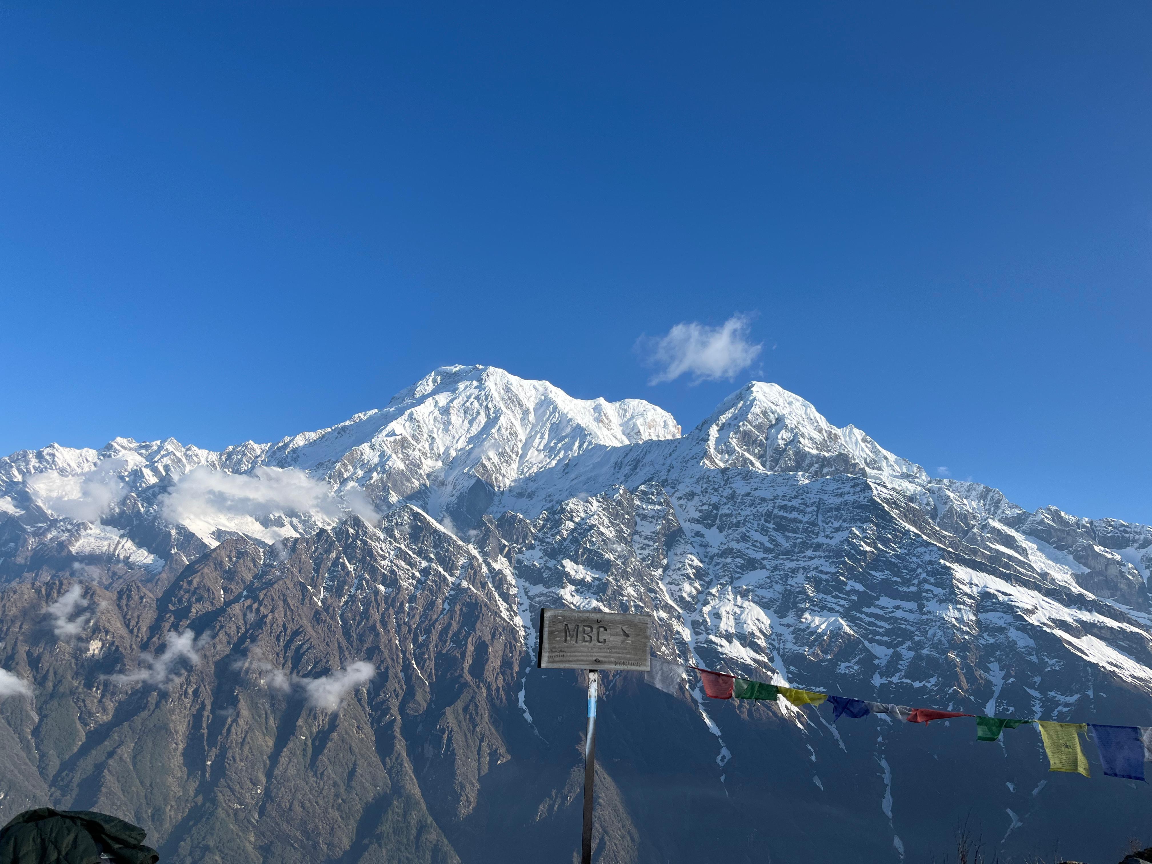 Snow capped mountain under a clear blue sky, with colorful prayer flags and a wooden sign labeled "MBC" in the foreground.