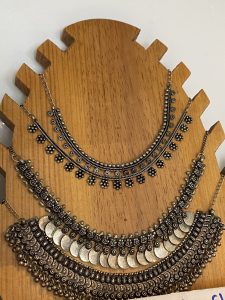 Three oxidized silver statement necklaces displayed on a wooden bust stand. The top two feature delicate floral charm chains, while the bottom is a wide bib-style necklace with coin pendants and intricate engraved motifs in traditional Indian style.
