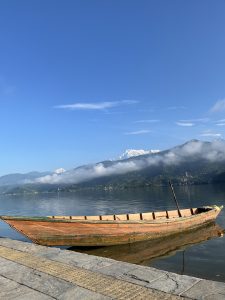 A wooden boat rests on a calm lake with a backdrop of misty mountains and snow-capped peaks under a clear blue sky.