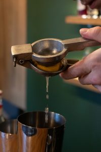 Close-up of a bartender hands squeezing fresh lemon juice through a metal citrus press into a cocktail shaker, with a green bar background.
