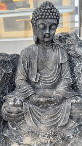 Ancient stone Buddha statue with intricate robe details sits serenely, eyes closed, hands in lap. Warm light behind creates a peaceful, contemplative mood.
