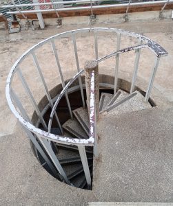 An outdoor concrete circular spiral staircase leading downward, enclosed by a weathered white metal safety railing. 