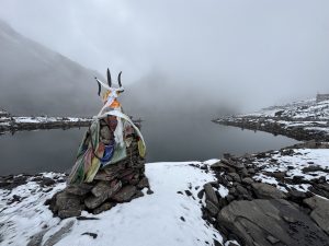 A wide-angle landscape shot of a stone cairn topped with a metal Trishul (trident) wrapped in white and colored ritual cloths. The cairn stands on a snowy, rocky shore next to a still, dark alpine lake. 
