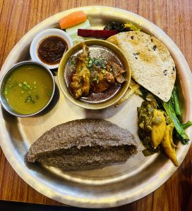 
A stainless steel plate with Indian cuisine: chicken curry, dal, vegetables, papad, millet bread, chutney, and salad. Rich colors reflect a traditional nepali meal.