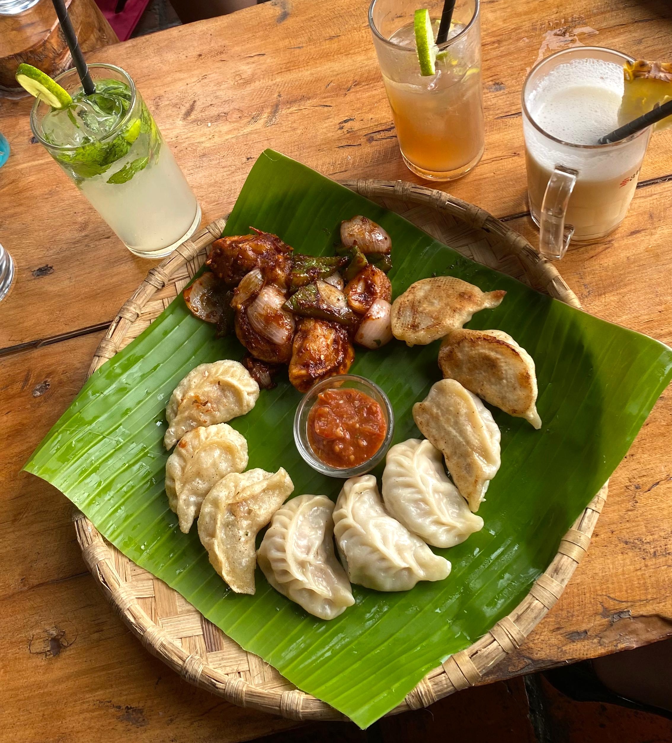 A round woven platter lined with a green banana leaf showcases a ring of pan-fried momos, a side of chili-sautéed meat with onions and bell peppers, and a central bowl of red tomato chutney, accompanied by three refreshing iced drinks on a wooden table.