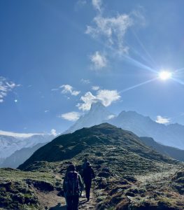 Hikers walk up a grassy mountain path under a bright sun with a snow-capped peak and blue sky in the background. Wispy clouds surround the mountain’s summit.