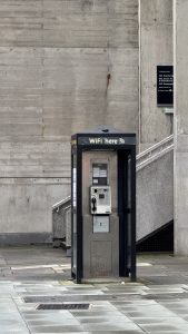 A vintage black payphone booth on a tiled sidewalk displays a “WiFi here” sign, blending old and new. Stairs rise to the left, while a textured concrete wall fills the background.
