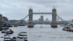 London’s Tower Bridge spans the Thames on a cloudy day, its stone towers and blue suspension cables standing out against a gray sky. Boats and barges float on the river below, with modern buildings visible in the background.
