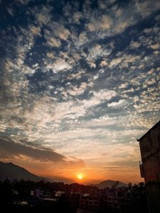 Dramatic sunset over a cityscape, with a glowing orange sun setting between silhouetted mountains under a sky scattered with textured clouds.