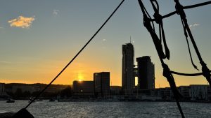 A waterfront cityscape at sunset, framed by the silhouetted ropes and rigging of a nearby boat. Modern buildings rise against a warm orange and yellow horizon, their colors reflected in the calm water below a softly clouded sky.
