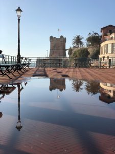 The promenade in Nervi (Genoa) with a puddle of water in the foreground, where you can see reflections of a lamppost and a castle tower with the Italian flag.