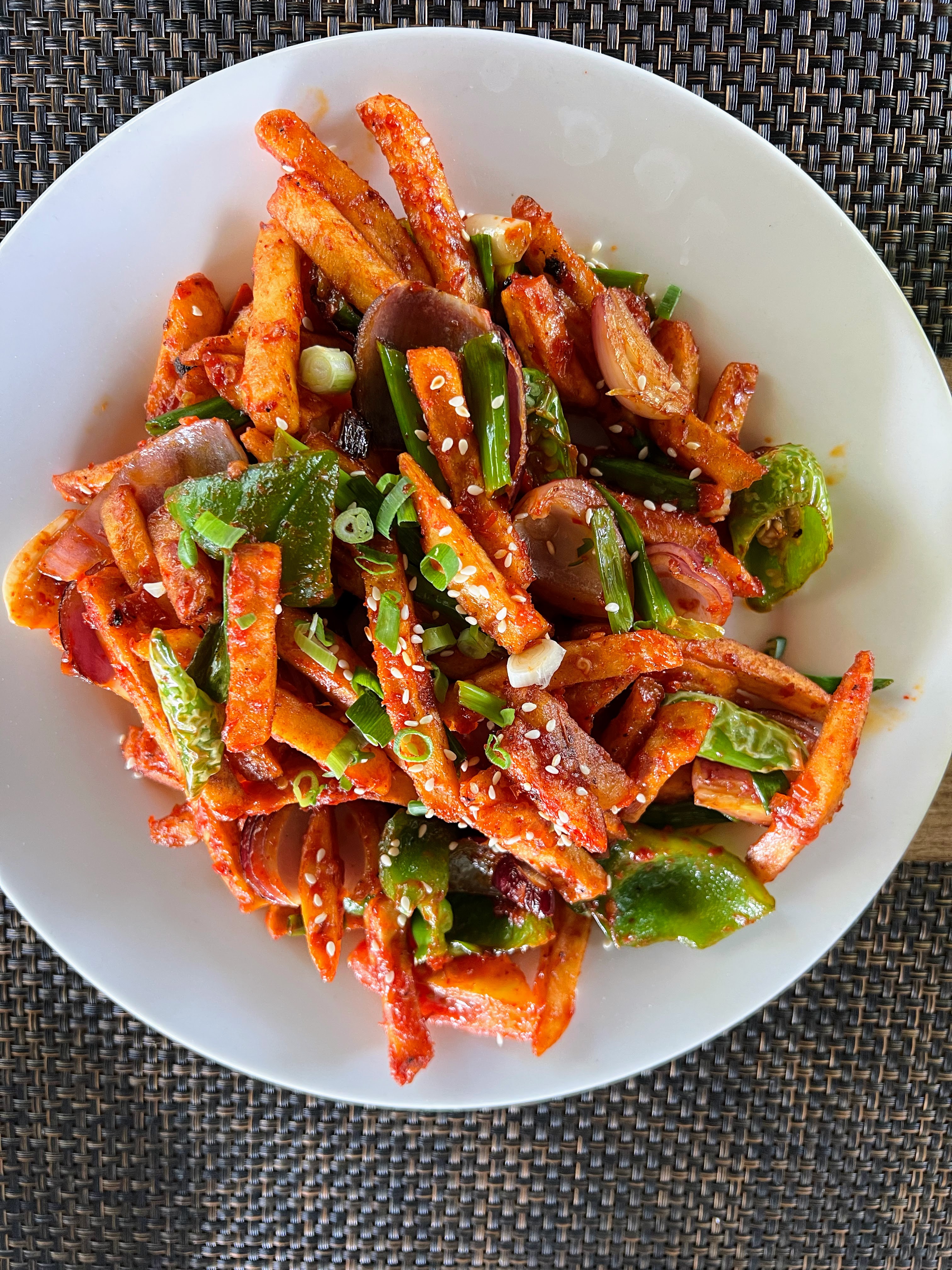 A top-down shot of a white plate filled with spicy, seasoned potato fries tossed with green peppers, onions, and sesame seeds.