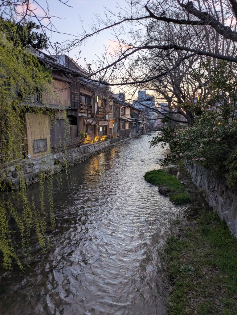 Peaceful evening view of the Shirakawa Canal in Kyoto’s historic Gion district, with traditional wooden machiya houses lining the waterfront. Willow branches, stone embankments, and softly illuminated streets create a serene atmosphere in Japan’s famous geisha quarter.