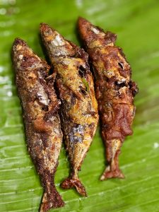 Three crispy fried Indian Mackerel are served on a fresh green banana leaf, showcasing the rich colors and textures of a traditional Kerala dish. The golden-brown crust and rustic presentation make the meal look both simple and delicious. Captured in Perumanna, Kozhikode.