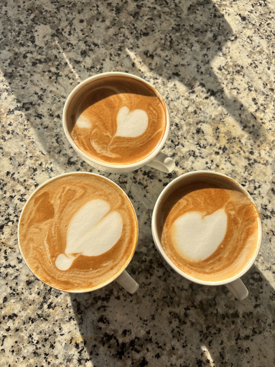 Three lattes with heart foam art on a speckled stone counter in warm morning sun.