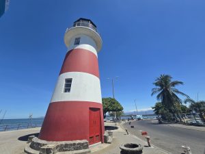 A red-and-white lighthouse stands beside a waterfront road lined with palm trees. The blue ocean and distant mountains are visible behind it under a clear blue sky.