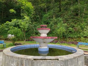 A weathered two-tiered concrete fountain with red-rimmed basins set in a circular blue-tiled pool, surrounded by blue park benches, purple ornamental plants, and a lush green forested hillside, Begnas Lake area, Nepal.