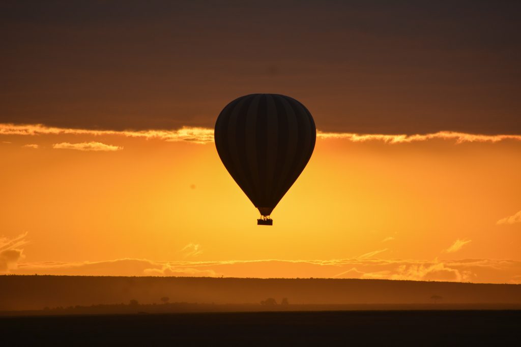 A hot air balloon floats over the Masai Mara Kenya landscape during sunset, silhouetted against a glowing orange sky. The warm evening light, distant horizon, and dramatic clouds create a peaceful safari atmosphere.