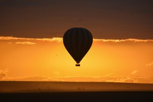 A hot air balloon floats over the Masai Mara Kenya landscape during sunset, silhouetted against a glowing orange sky. The warm evening light, distant horizon, and dramatic clouds create a peaceful safari atmosphere.
