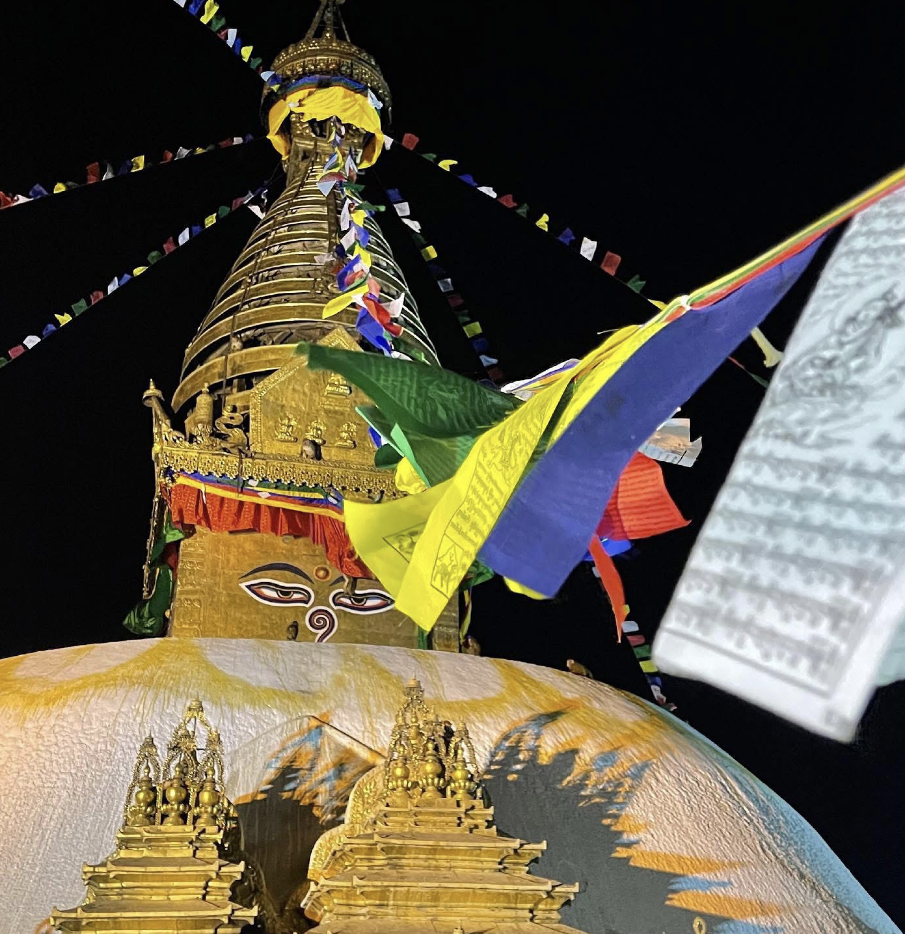 Golden stupa with Buddha eyes at night, adorned with vibrant prayer flags. The scene conveys peace and spirituality against a dark sky.