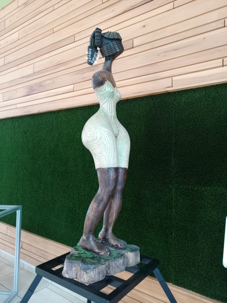 A tall, slender wooden sculpture of a stylized female figure featuring dark wood tones on the limbs, an elongated neck, a textured black hairstyle, and a light-colored dress detailed with faint green wavy lines.