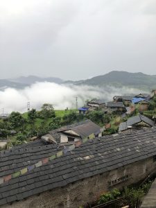 A high-angle view of a traditional Nepalese hillside village with stone-tiled and tin roofs. Colorful Buddhist prayer flags are strung across the rooftops, overlooking lush green terraces and deep valleys blanketed in thick, rolling white clouds under an overcast sky. 