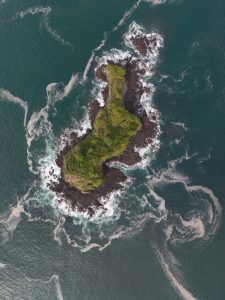 An aerial view of a small rocky island covered with green vegetation, surrounded by deep blue ocean. White waves and sea foam swirl around the dark rocks, creating patterns across the water.