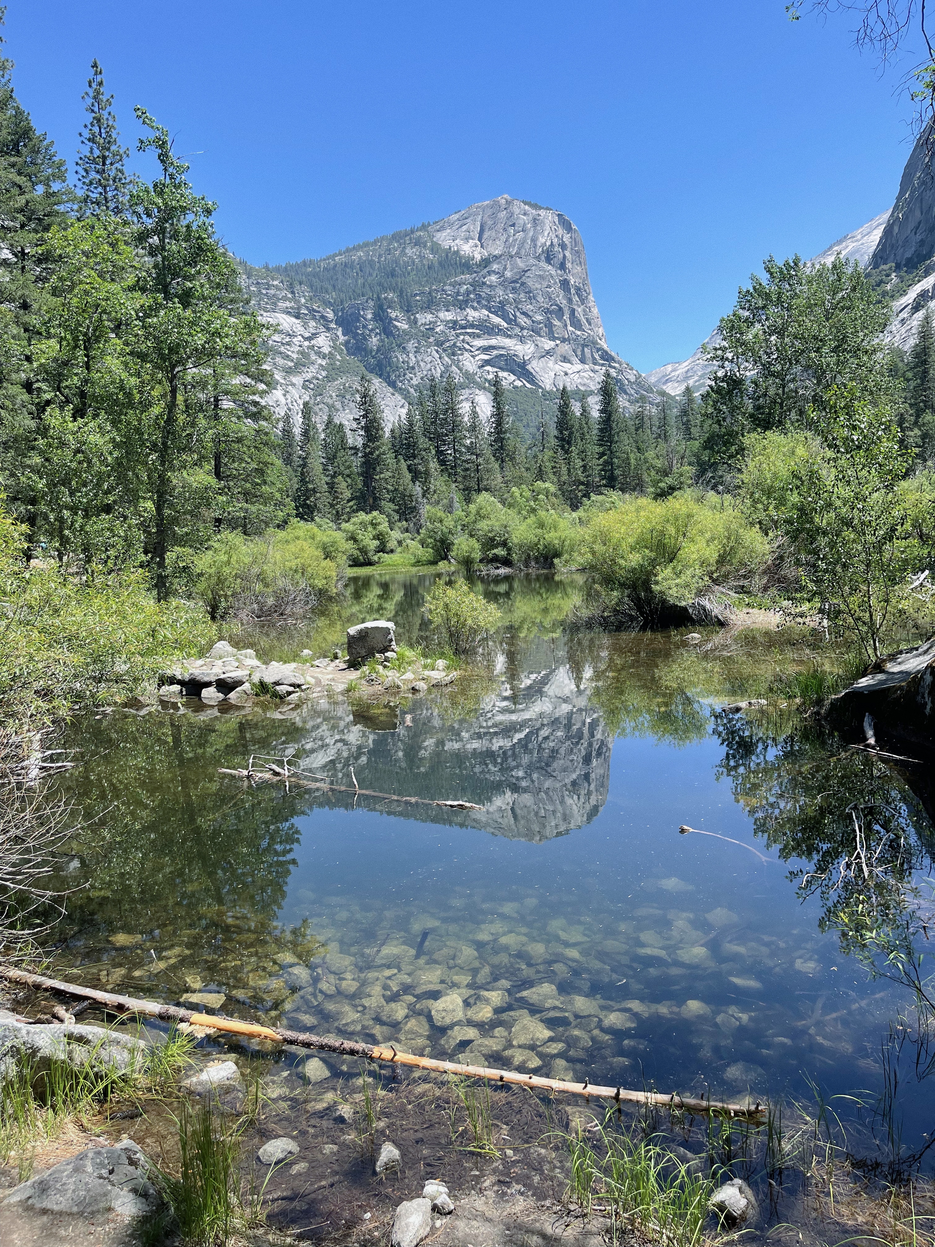 A serene landscape of a clear pond reflecting a towering mountain and lush greenery under a bright blue sky