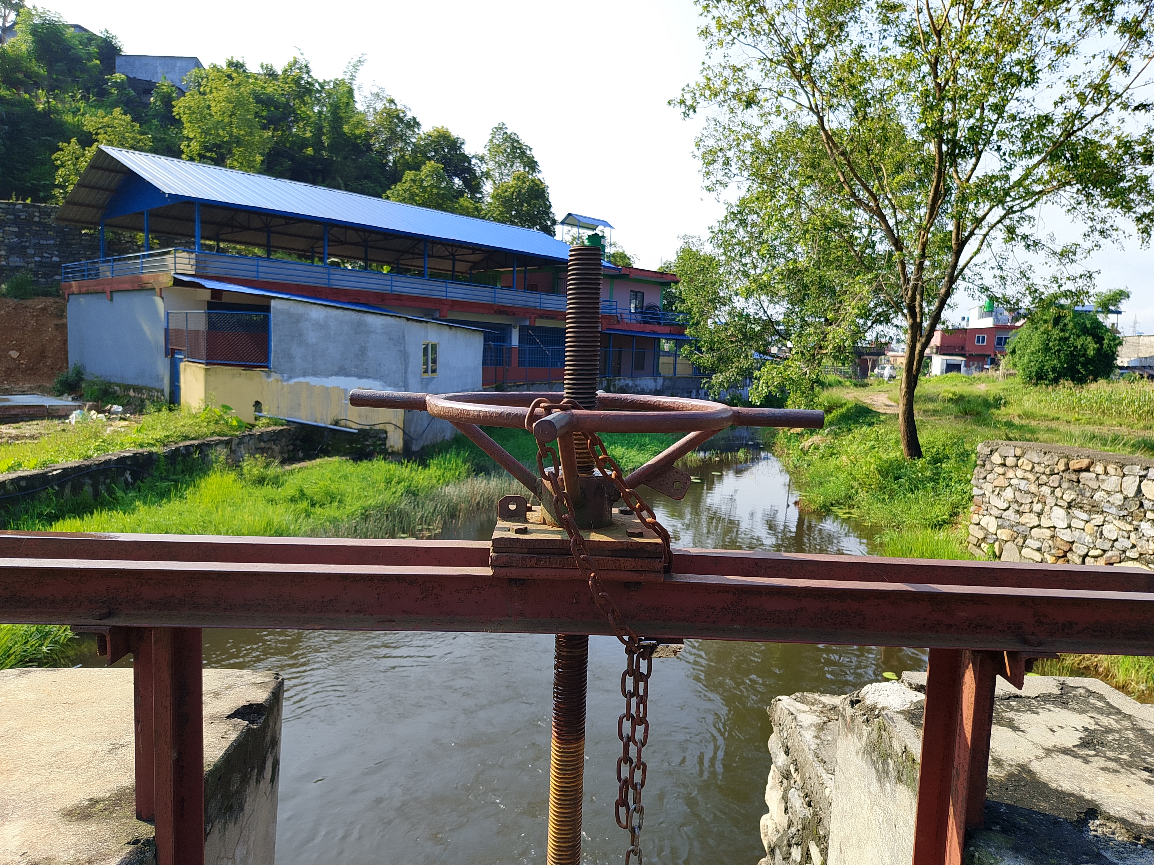 A rusty hand-operated sluice gate mechanism over a calm canal with lush greenery behind.