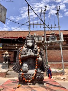 A dark stone Garuda statue in a praying namaste posture, decorated with marigold garlands, standing on a plinth in a Kathmandu temple courtyard with utility poles and brick buildings in the background. 