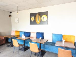 Cafe interior with a long blue bench, small tables, light brown chairs, and abstract gold wall art on a white wall.
