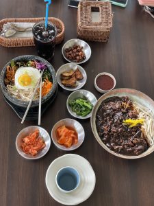 Top-down view of a wooden table with Korean dishes, including bibimbap, jajangmyeon, kimchi side dishes, and an iced drink with a straw.