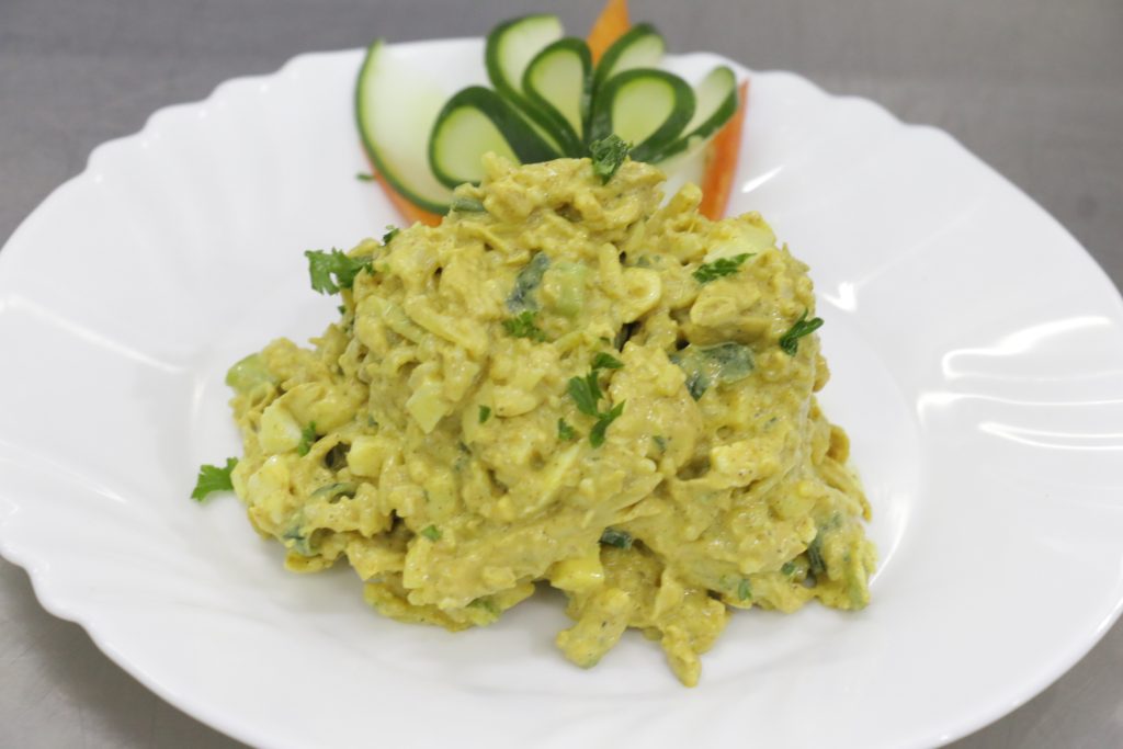 Creamy yellow salad with chopped vegetables and herbs served on a white plate, garnished with parsley and a decorative fan of thin cucumber slices in the background.