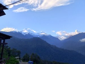 Snow-capped Himalayan peaks at dusk with forested ridges and prayer flags below

