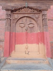 Historic wooden door with intricate carvings and large eye motifs, set in a brick wall with faded red paint. 