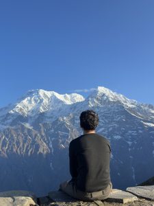 A person in a dark sweater sits quietly on a stone ledge, seen from behind, contemplating a magnificent panoramic view of snow-covered Himalayan peaks under a clear blue sky. 