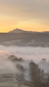 A hilltop structure with a tall steeple silhouettes against a warm dawn sky, while layers of soft fog roll across fields and a small village below. Trees and rooftops emerge faintly from the mist, creating a tranquil, atmospheric scene.
