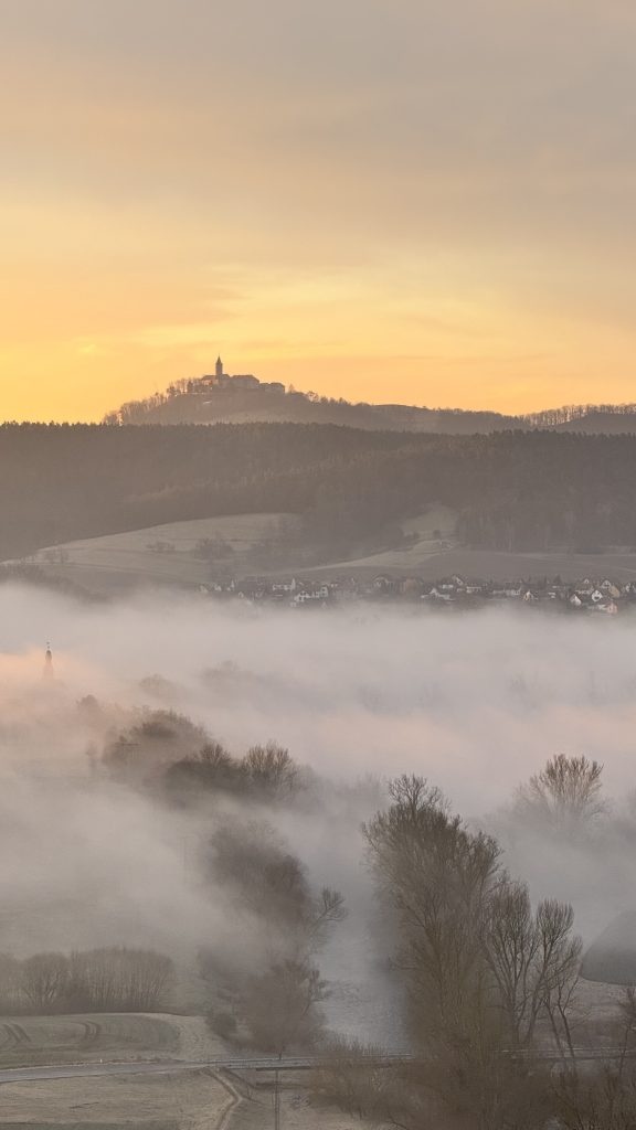 A hilltop structure with a tall steeple silhouettes against a warm dawn sky, while layers of soft fog roll across fields and a small village below. Trees and rooftops emerge faintly from the mist, creating a tranquil, atmospheric scene.