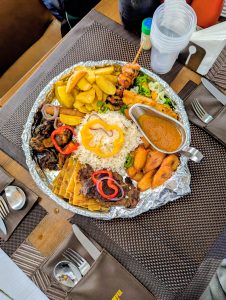 A large platter with white rice, grilled chicken skewers, grilled meat, fried plantains, potato wedges, vegetables, salad, and a dipping sauce, served on a table with cutlery.
