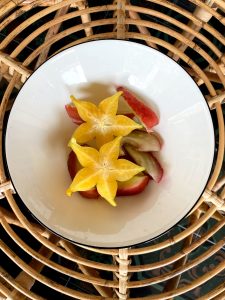 A white bowl with vibrant yellow starfruit slices and red apple wedges sits on a woven rattan table.
