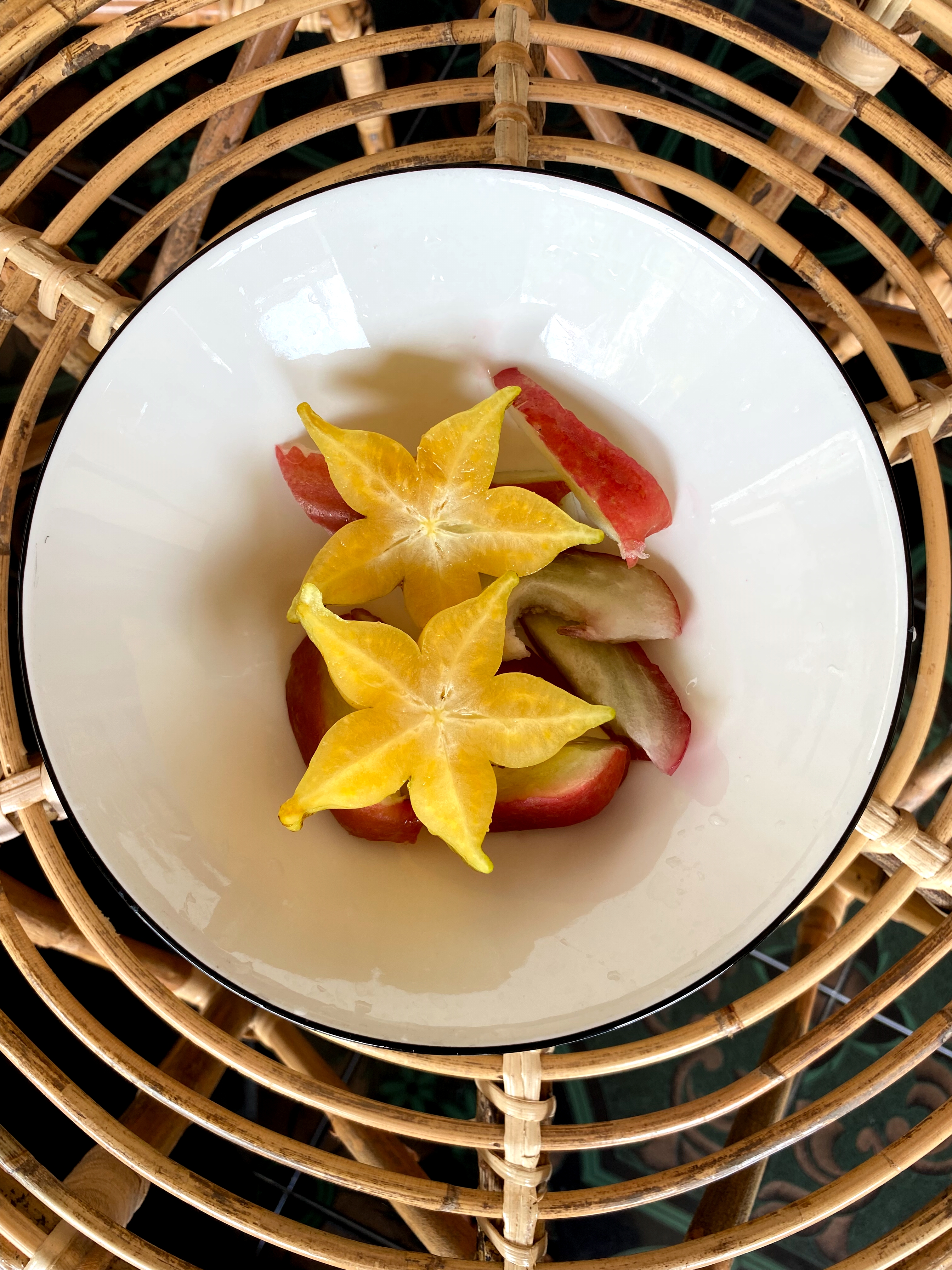 A white bowl with vibrant yellow starfruit slices and red apple wedges sits on a woven rattan table.