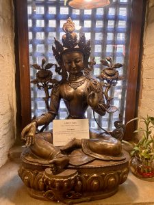 A polished bronze Green Tara statue seated on a lotus throne with a descriptive placard, flanked by lotus flowers, displayed in a traditional Nepali latticed window niche. 