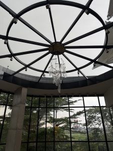 
A circular glass ceiling with a metal frame supports a transparent abstract sculpture. Large windows below reveal lush greenery and a distant lighthouse.