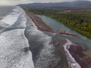 An aerial view of a river mouth meeting the ocean along a wide coastline. White waves roll onto dark sand, while turquoise river water curves beside dense green forest and distant mountains.