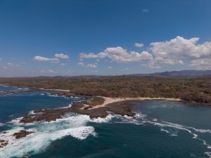 An aerial view of a rocky coastline with small sandy beaches and deep blue water. White waves break against the rocks, while dry forest, distant hills, and scattered clouds fill the background.