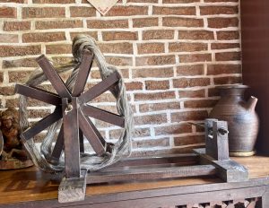 A small traditional wooden spinning wheel with natural grey yarn wound around it, displayed beside a dark ceramic jug on a wooden shelf against an exposed brick wall. 
