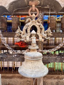 An ornate silver-toned temple bell with naga (serpent) crown motif hanging on a metal chain, with large brass prayer wheels and a blue tarpaulin visible in the background, Nepal. 