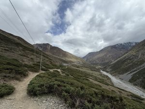 A high altitude trekking trail winding through a barren mountain valley with a glacial river and a small remote village in the distance.