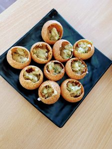 A top-down shot of a black square plate filled with ten crisp, hollow puri shells stuffed with a savory potato and herb mixture, placed on a light wooden table.