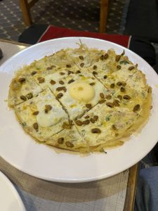 A top-down shot of a  rice crepe served on a round white plate. The crepe is topped with minced meat, scattered black-eyed beans, a drizzle of savory oil, and a sunny-side-up egg cooked perfectly in the center, all sliced neatly into triangles.